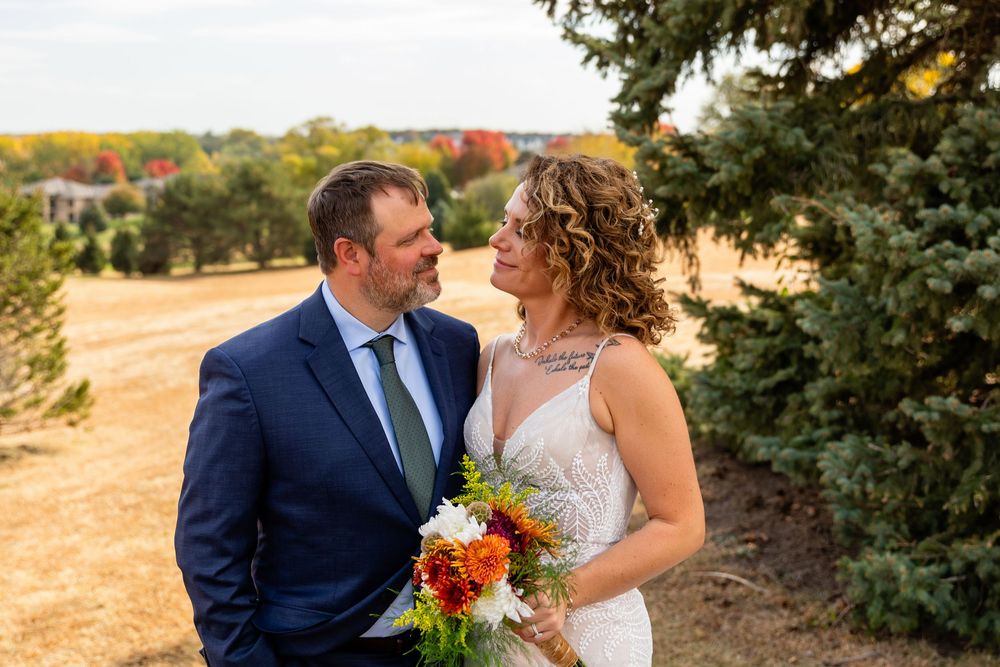 Bride holding autumn bouquet and groom in navy suit share a tender glance outdoors among fall foliage  at a Venue on the Ridge in Elkhorn NE
