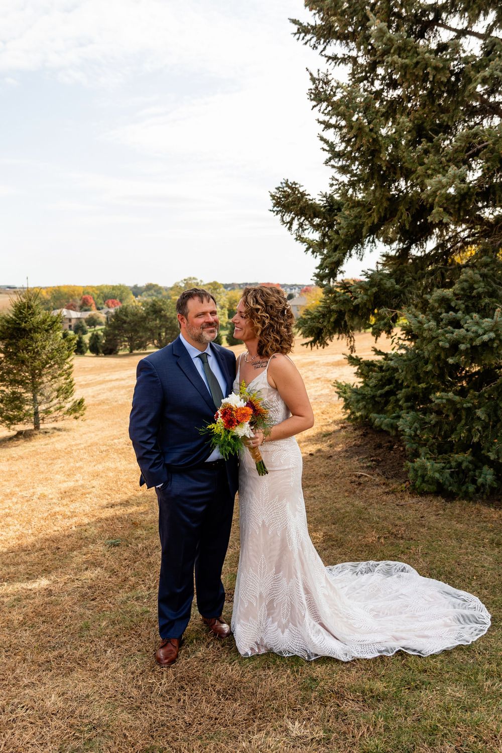 Bride and groom share a romantic glance while standing in a golden field beside an evergreen tree  at a Venue on the Ridge in Elkhorn, NE