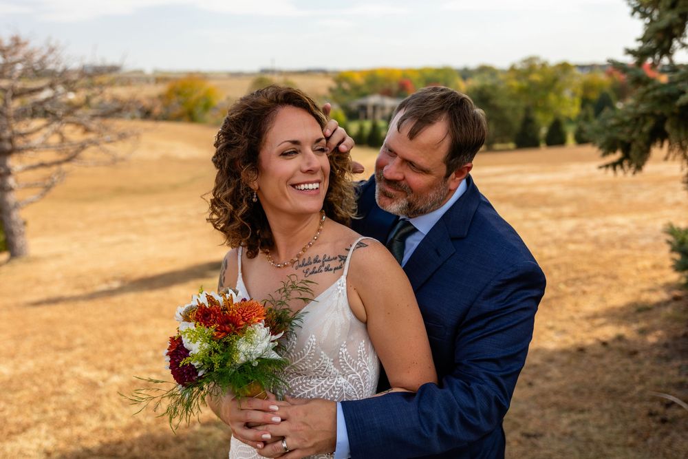 Smiling bride with bouquet and groom in navy suit share a tender moment outdoors during fall wedding  at a Venue on the Ridge in Elkhorn, NE