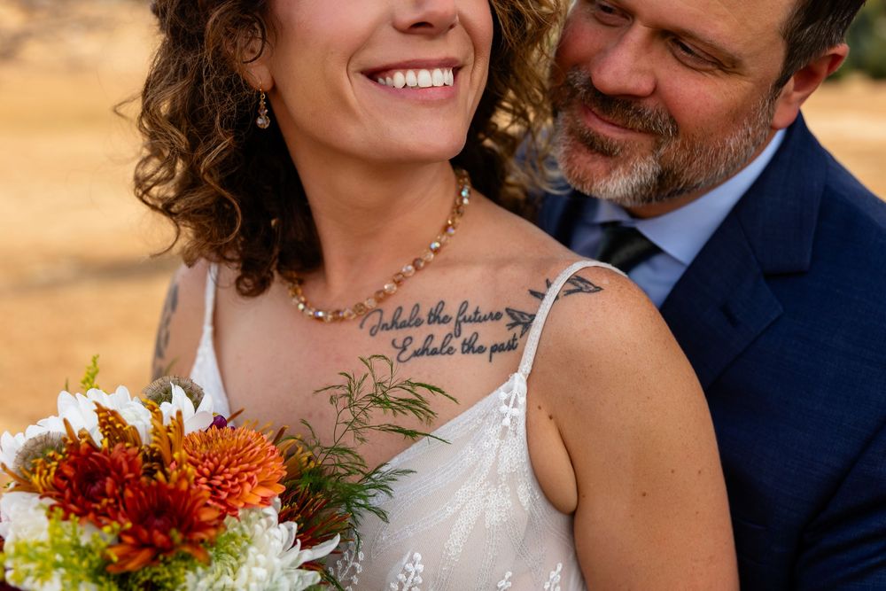 Smiling bride with curly hair holding orange and white bouquet embraced by groom in navy suit outdoors.