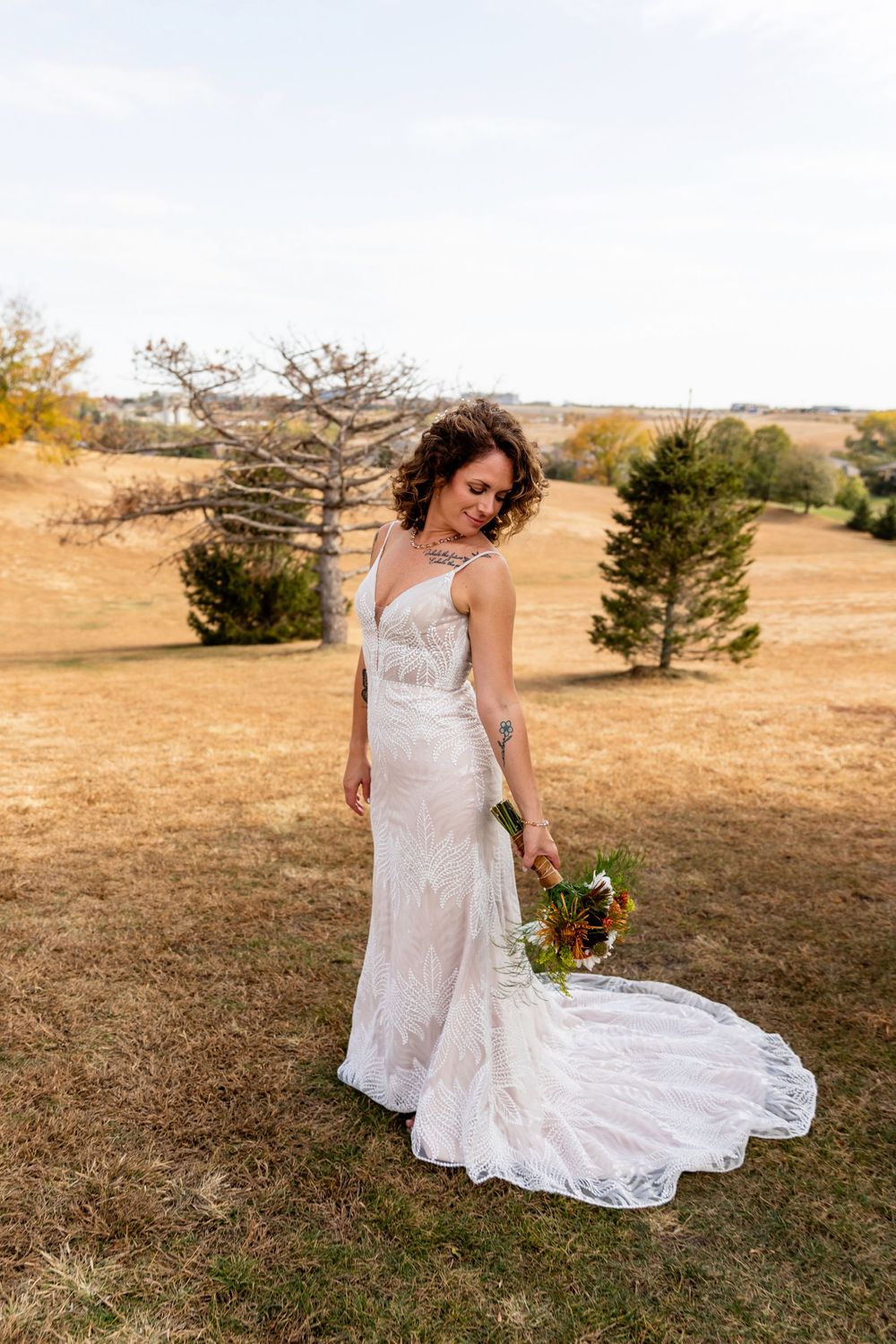 Bride in backless lace gown stands in golden field holding a colorful autumn bouquet on her wedding day  at a Venue on the Ridge in Elkhorn