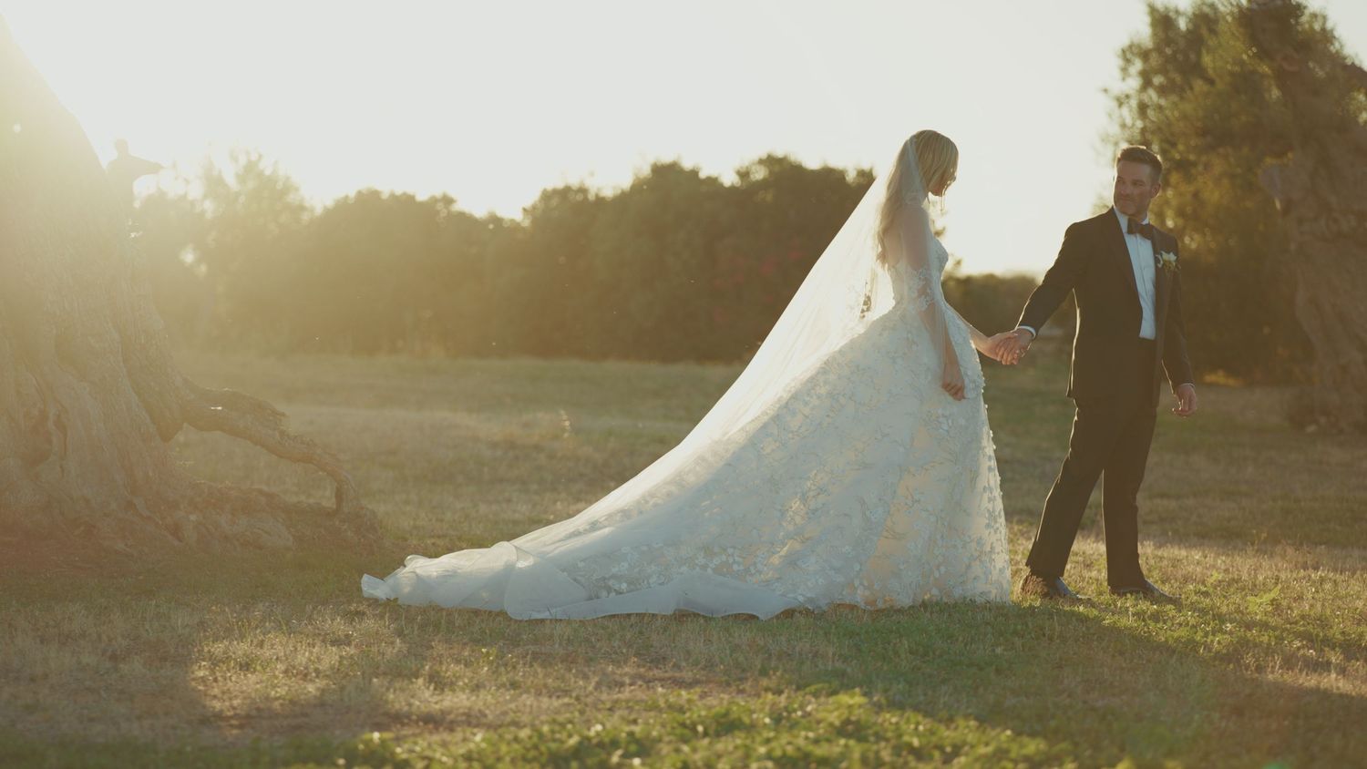 A bride in a flowing white lace gown and groom in tuxedo walk hand in hand at golden hour in a sunlit field.