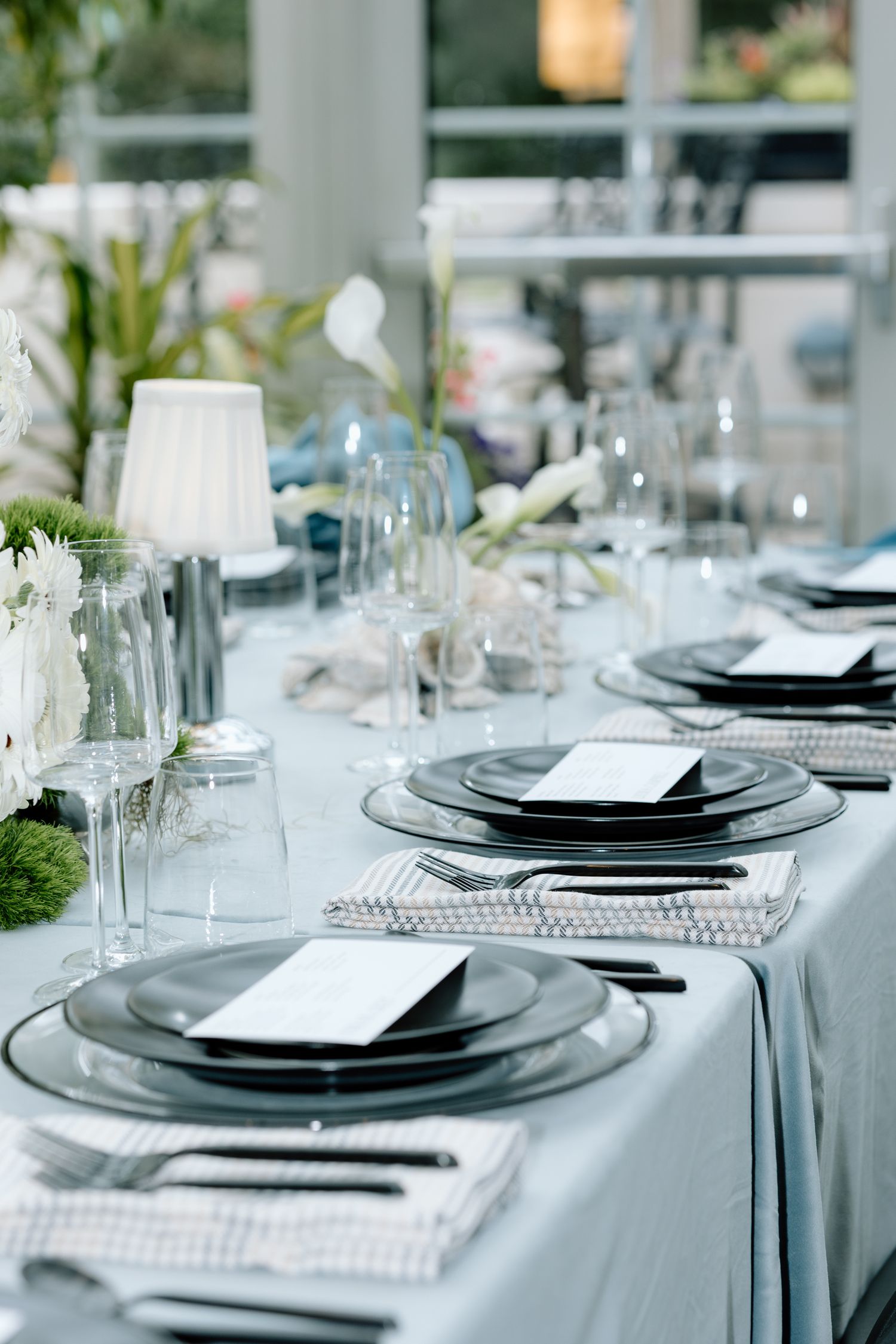 Elegant table setting with dark plates, crystal glasses, white floral centerpieces, and pearl napkin rings on gray linen.