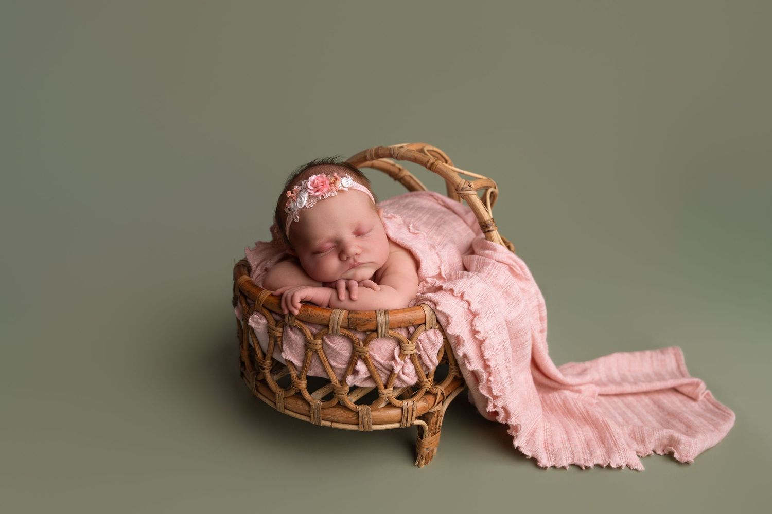 Newborn baby girl sleeping peacefully in a wicker basket with pink wrap and floral headband on green background.