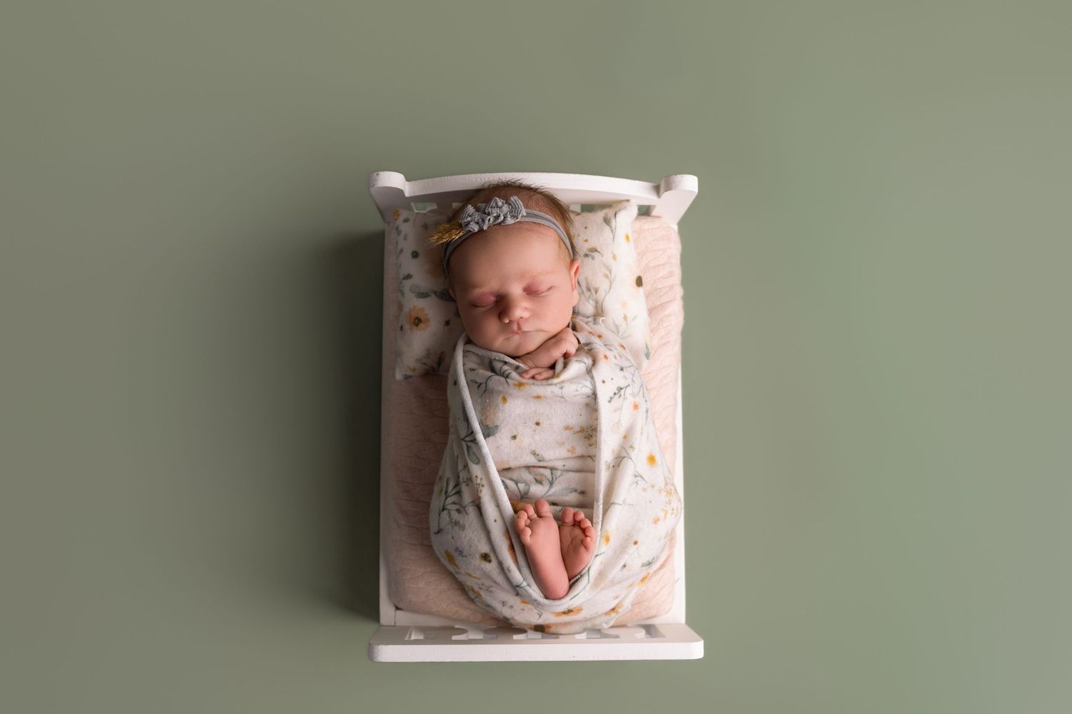 Newborn baby swaddled in floral wrap sleeping peacefully in a small white wooden bed against a sage green background.