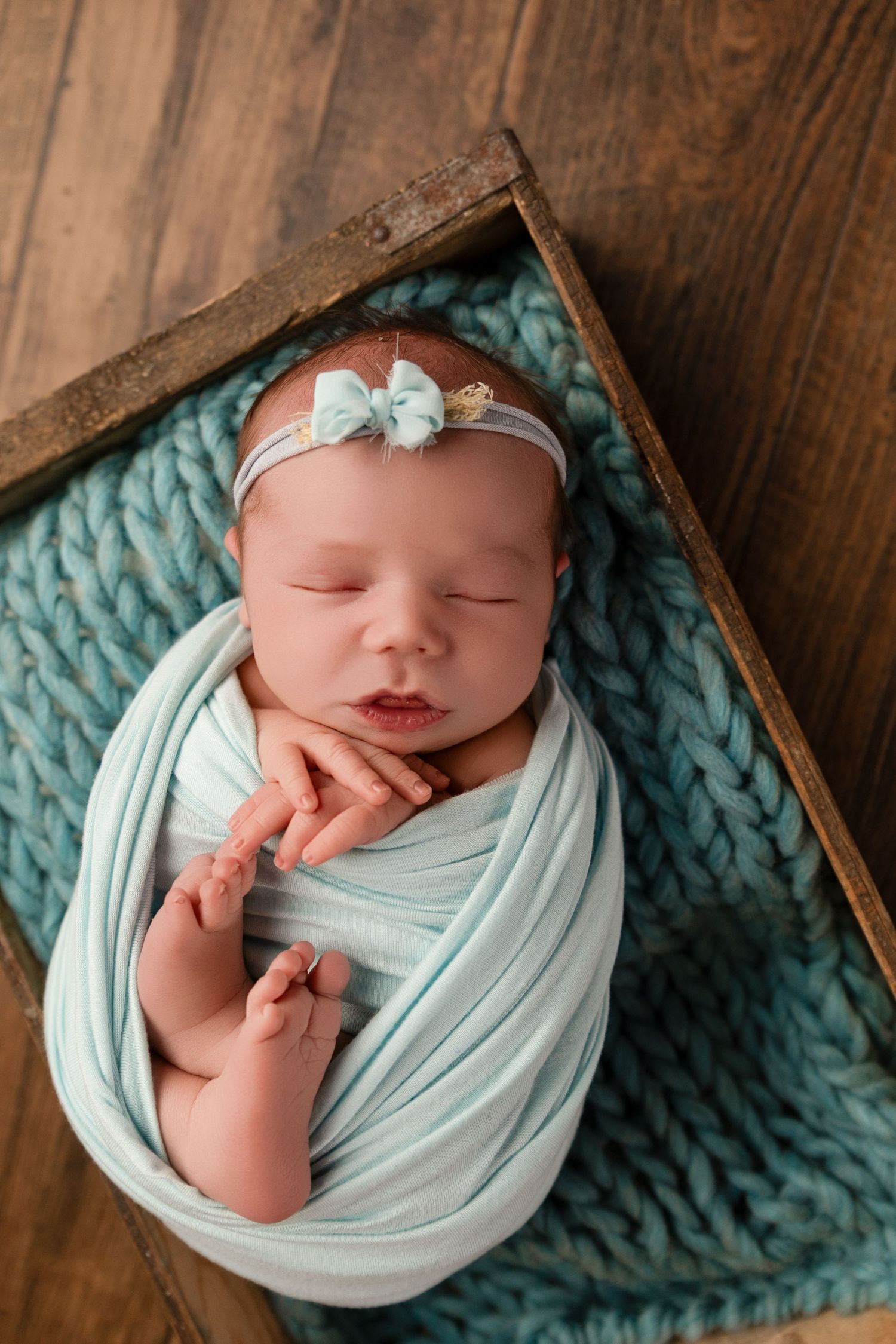 Newborn baby girl swaddled in teal wrap with bow headband sleeping peacefully in rustic wooden box.