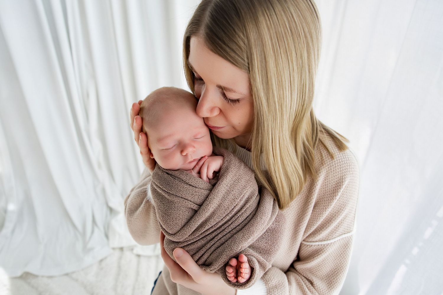 A mother tenderly kisses her sleeping newborn baby wrapped in a soft brown swaddle blanket.
