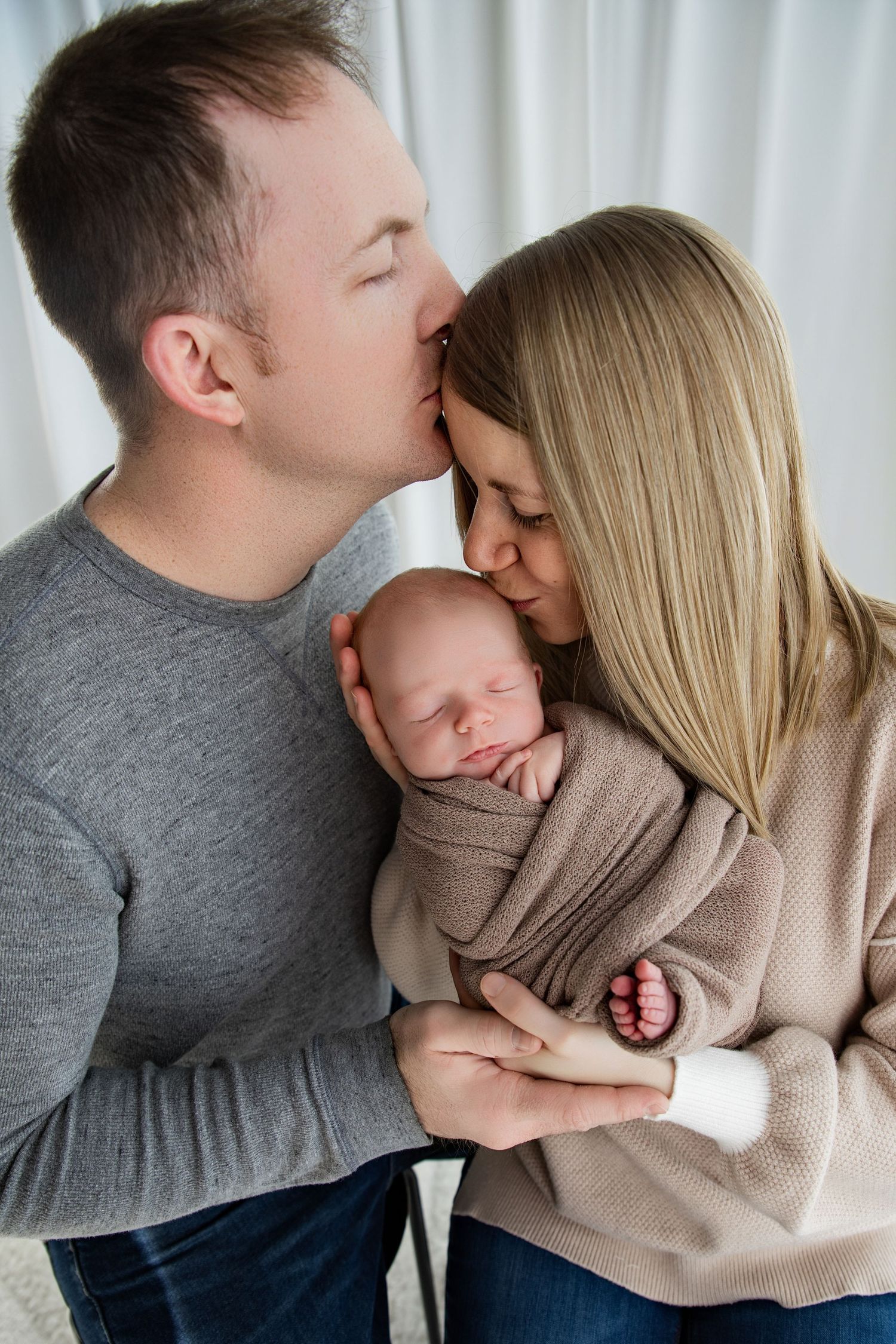 A couple tenderly kisses their sleeping newborn baby wrapped in a beige swaddle blanket.