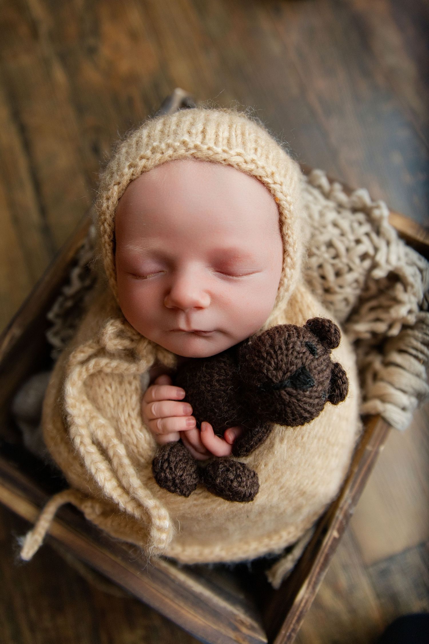 Sleeping newborn baby in cream knit bonnet and wrap, cuddling a small brown crocheted teddy bear in a wooden crate.
