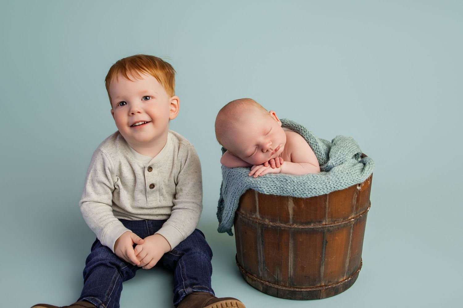 A smiling toddler with red hair sits beside a sleeping newborn in a rustic wooden bucket on a blue backdrop.
