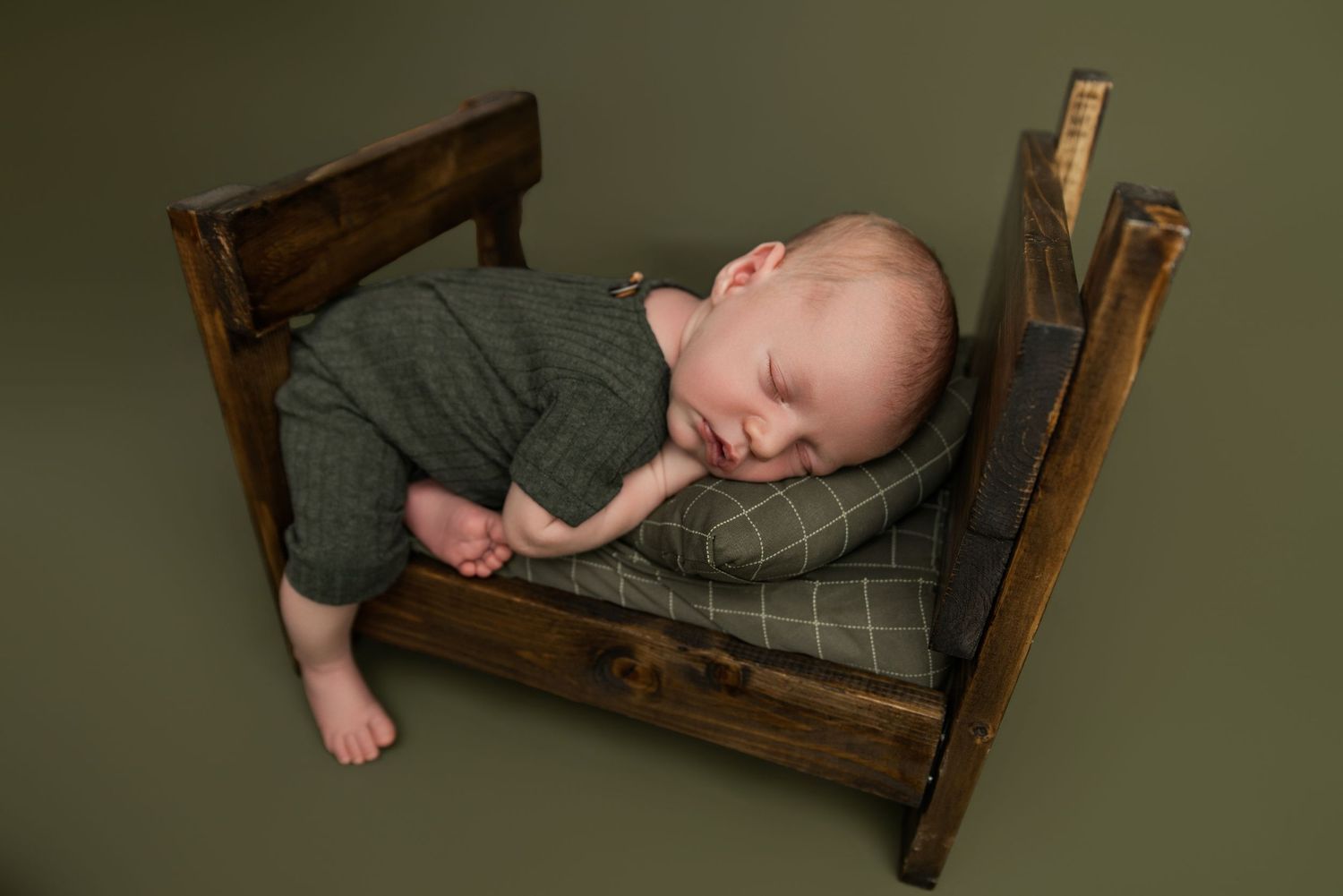 Newborn baby sleeping peacefully on a tiny wooden bed with green bedding, wearing a gray outfit, on a green background.