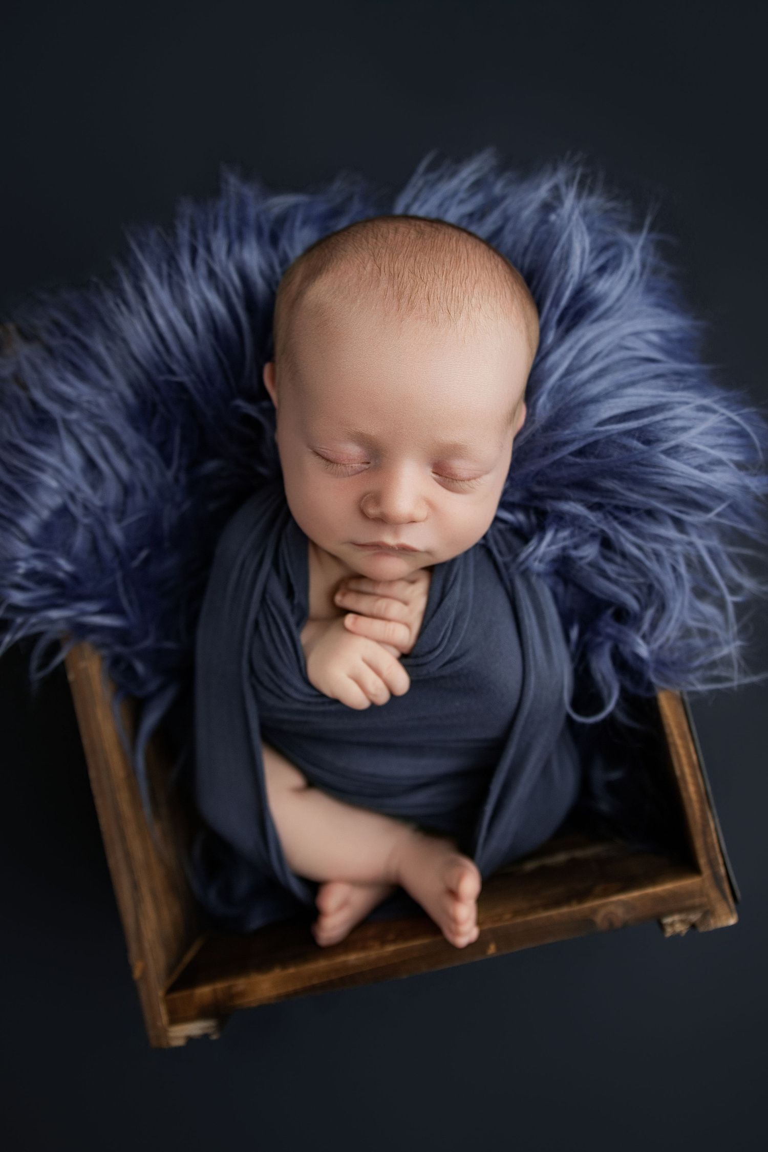 Sleeping newborn baby wrapped in blue swaddle cloth resting in rustic wooden box with fluffy blue fur blanket.