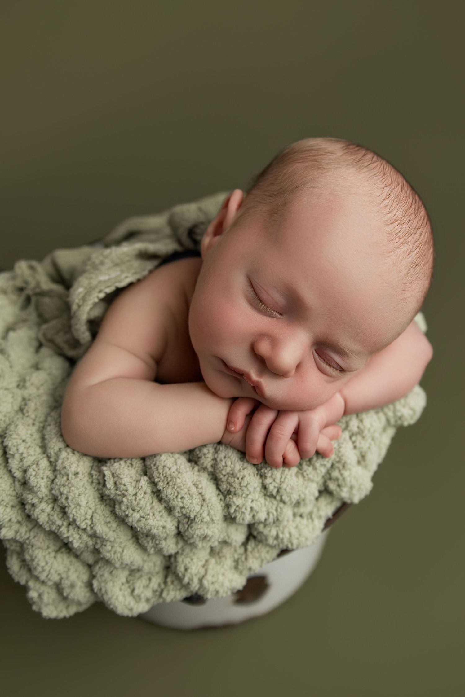 Sleeping newborn baby nestled in a white bucket with sage green knit blanket against olive green background.