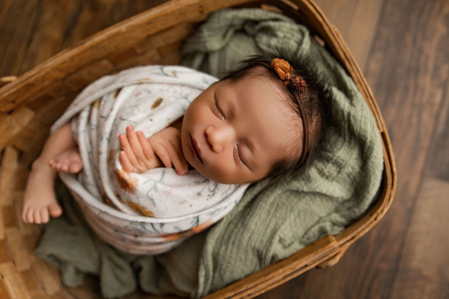 Sleeping newborn baby swaddled in white wrap, nestled in a wicker basket with green fabric and floral headband.
