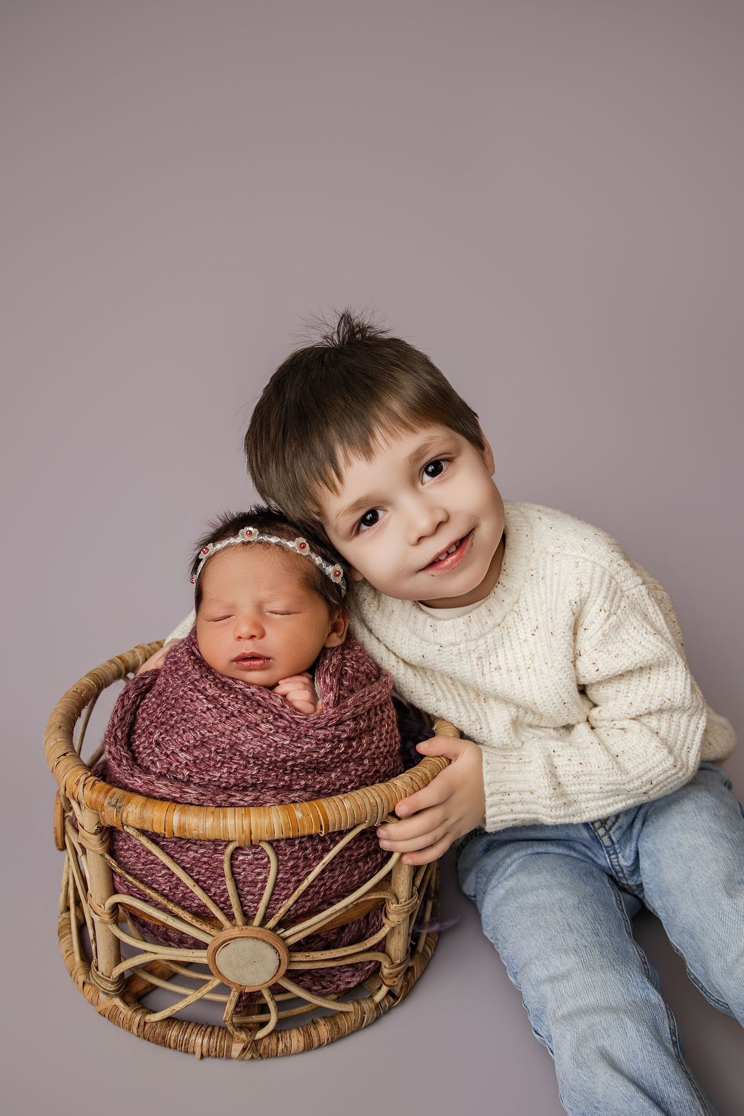Young boy in cream sweater proudly poses with sleeping newborn sibling wrapped in pink knit inside wicker basket.