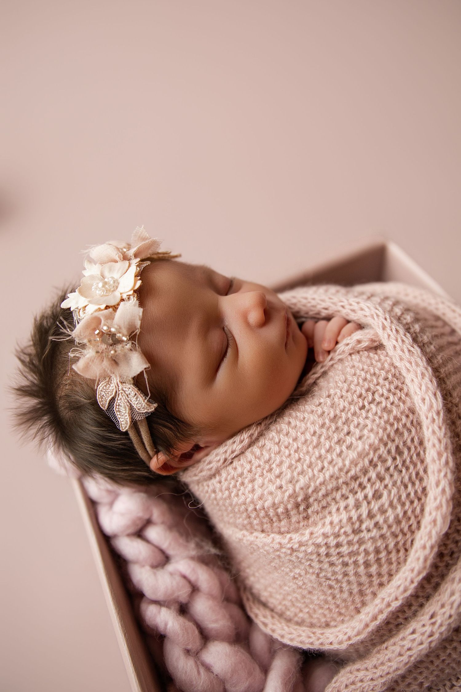 Newborn baby girl swaddled in pink knit wrap with floral headband, sleeping peacefully in a wooden box.