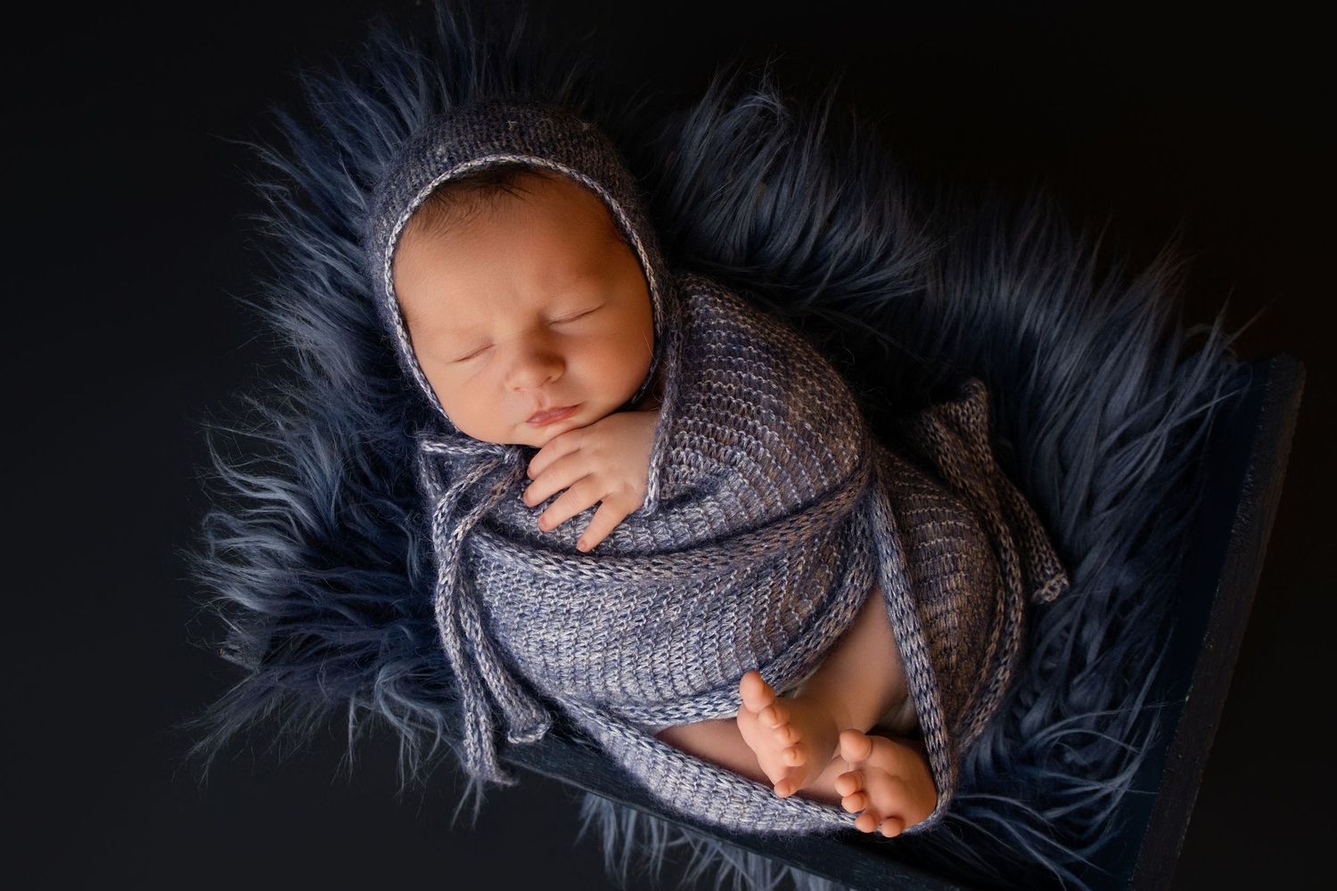 Newborn baby swaddled in blue knit wrap and bonnet, sleeping peacefully on fluffy blue fur blanket.