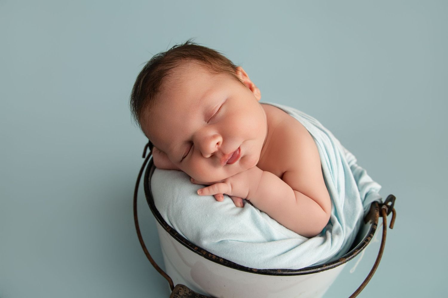 Newborn baby sleeping peacefully in a white bucket wrapped in a light blue swaddle against a blue background.