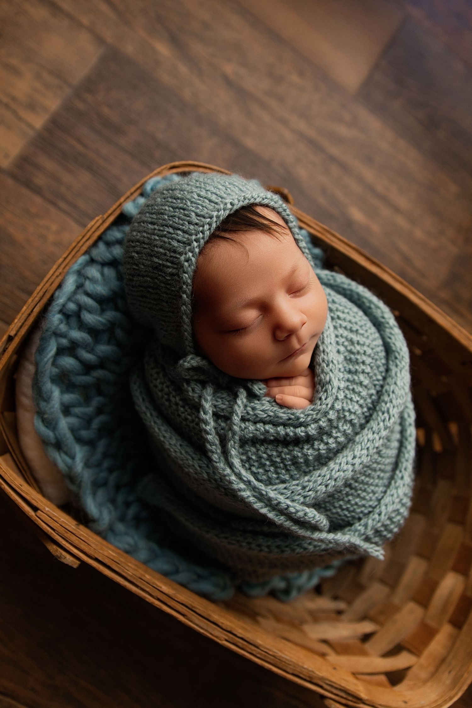 Sleeping newborn baby swaddled in a teal knit wrap and bonnet, nestled in a wicker basket on a wood floor.
