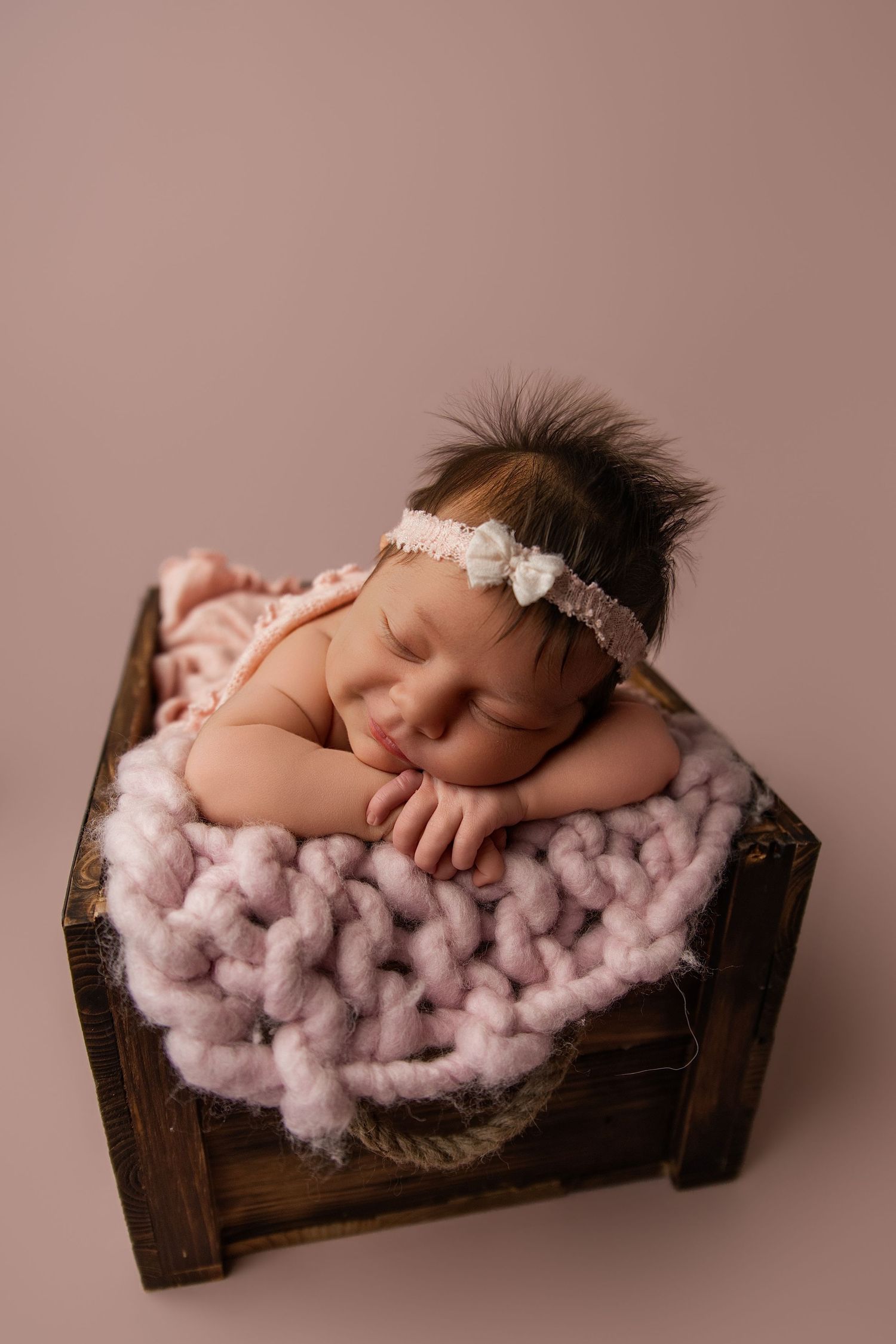 Sleeping newborn baby girl in wooden crate with pink knit blanket and floral headband on mauve background.