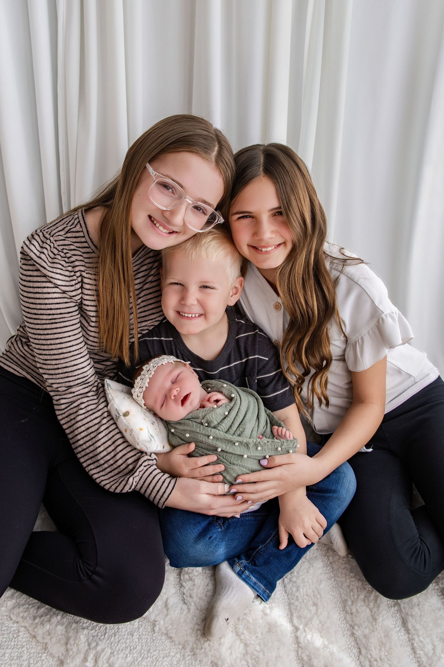 Three siblings smile together, two older girls and a boy holding a sleeping newborn baby wrapped in green.