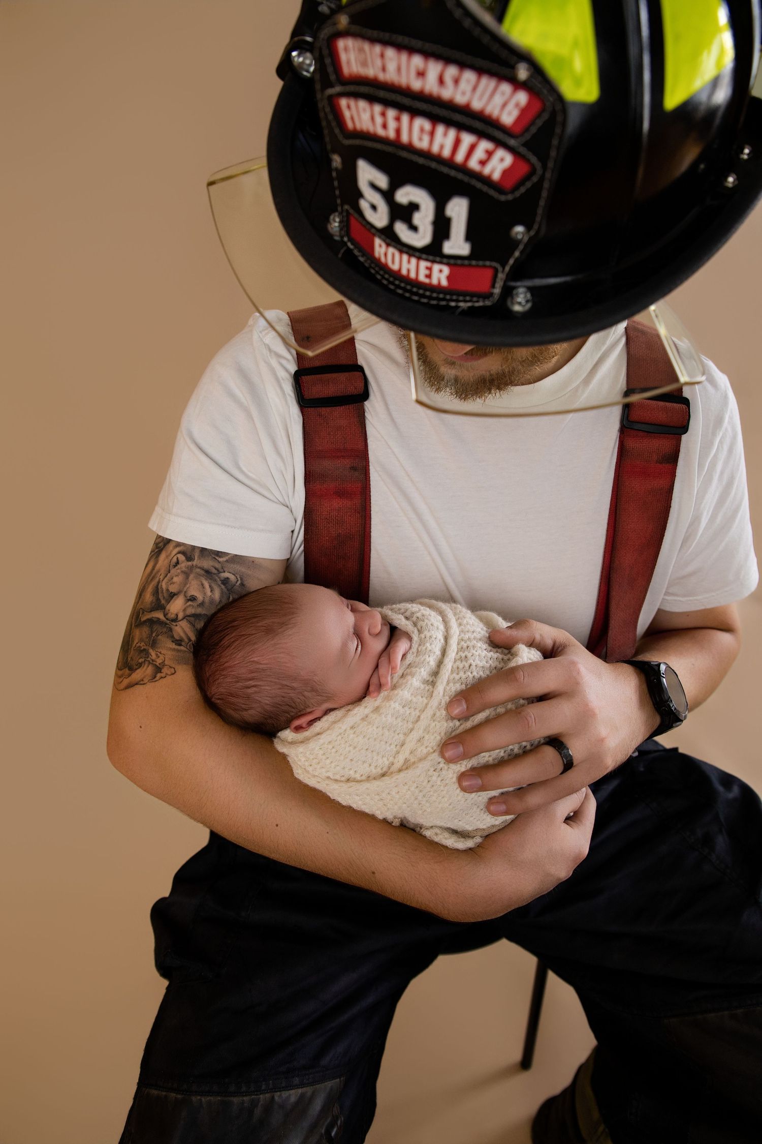 Fredericksburg firefighter 531 holding a swaddled newborn baby, wearing helmet and suspenders with tattoed arms.