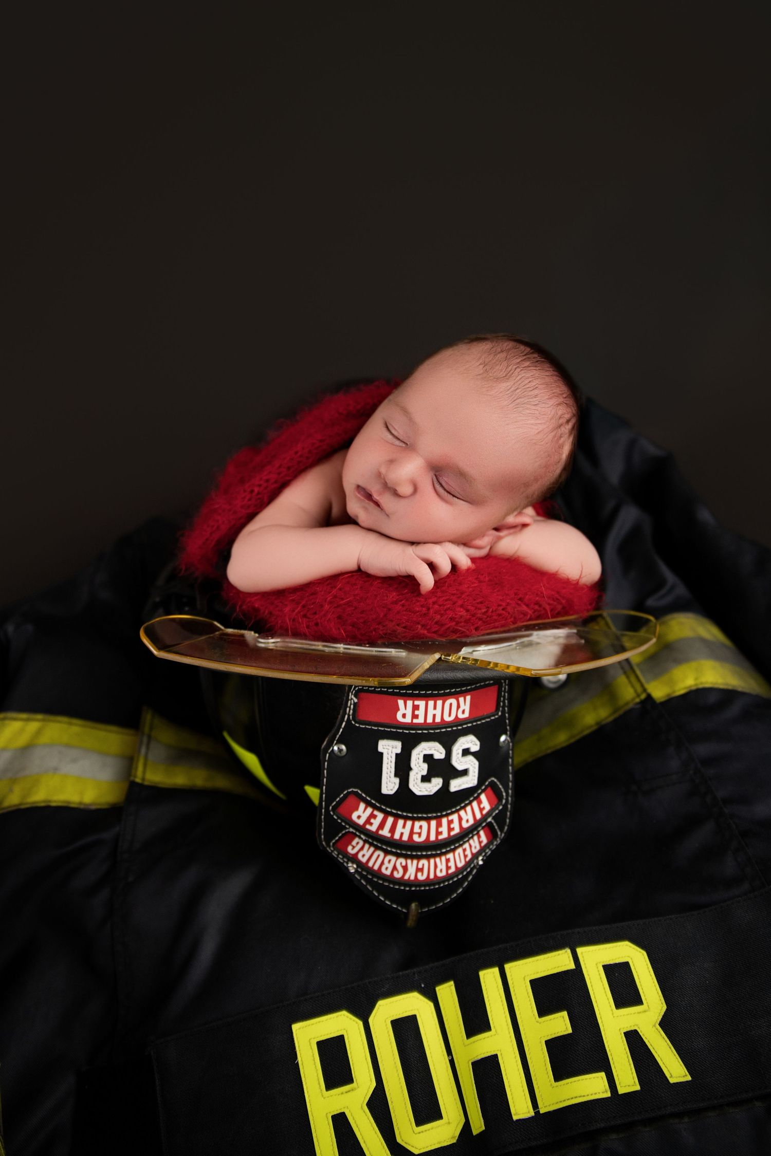Newborn baby sleeping peacefully on a firefighter helmet atop a Roher turnout gear jacket with red blanket.