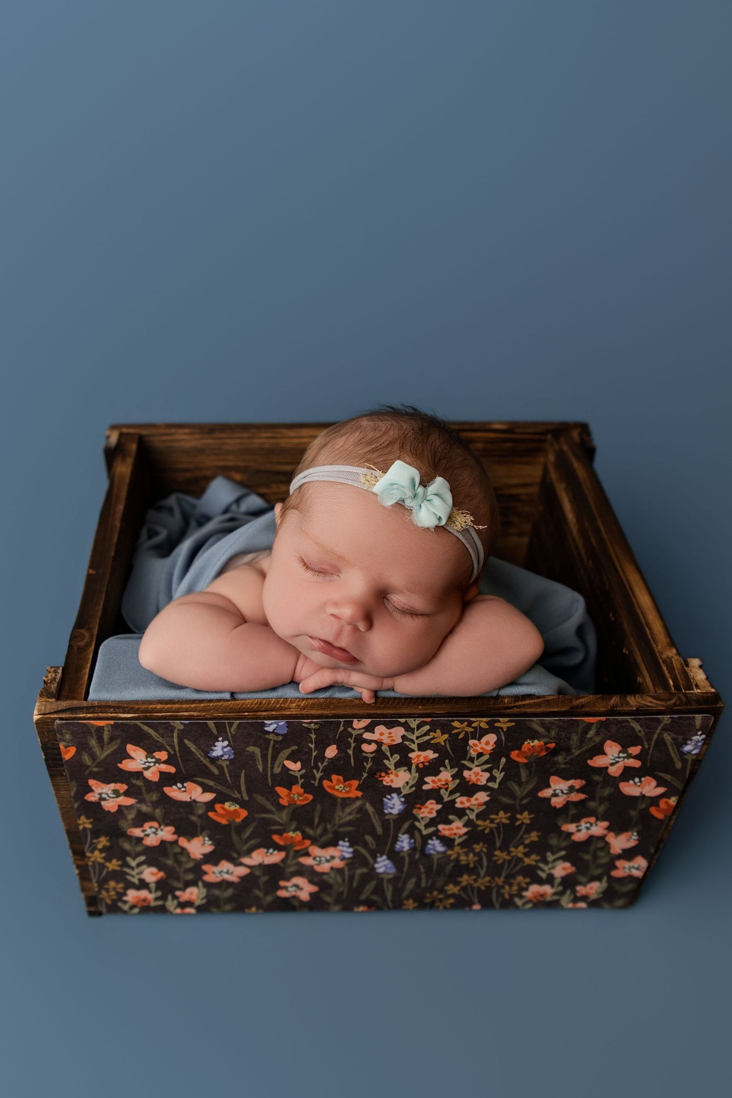 Sleeping newborn baby girl in floral wooden crate with blue blanket and mint flower headband on blue background.