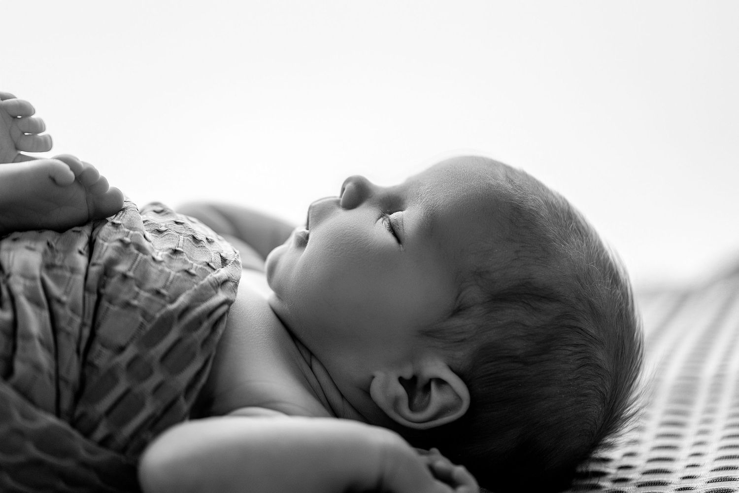 Black and white photo of a peaceful sleeping newborn baby lying on a textured surface wearing a patterned outfit.