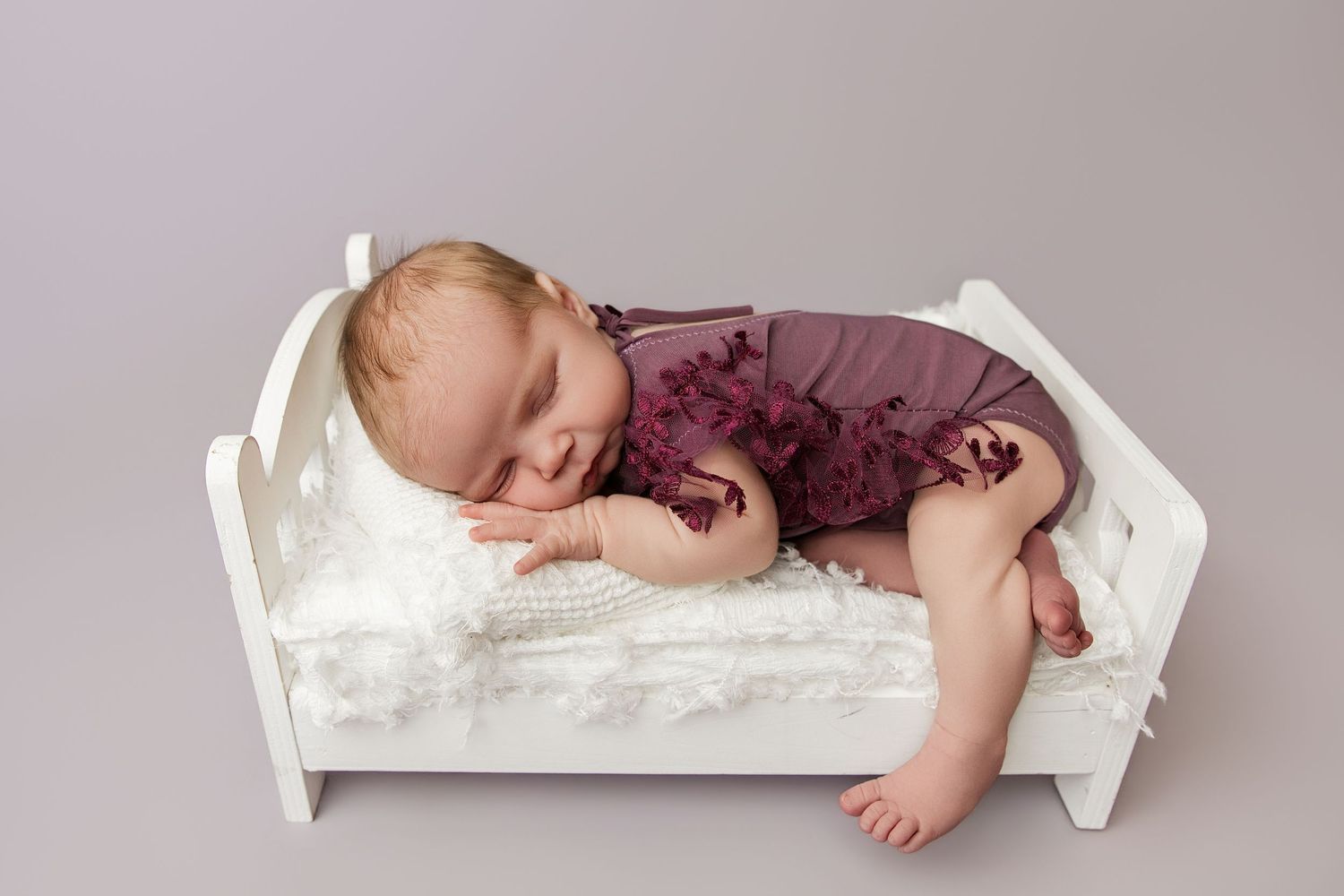 Sleeping newborn baby in purple romper posed on tiny white wooden bed with fluffy white blanket.