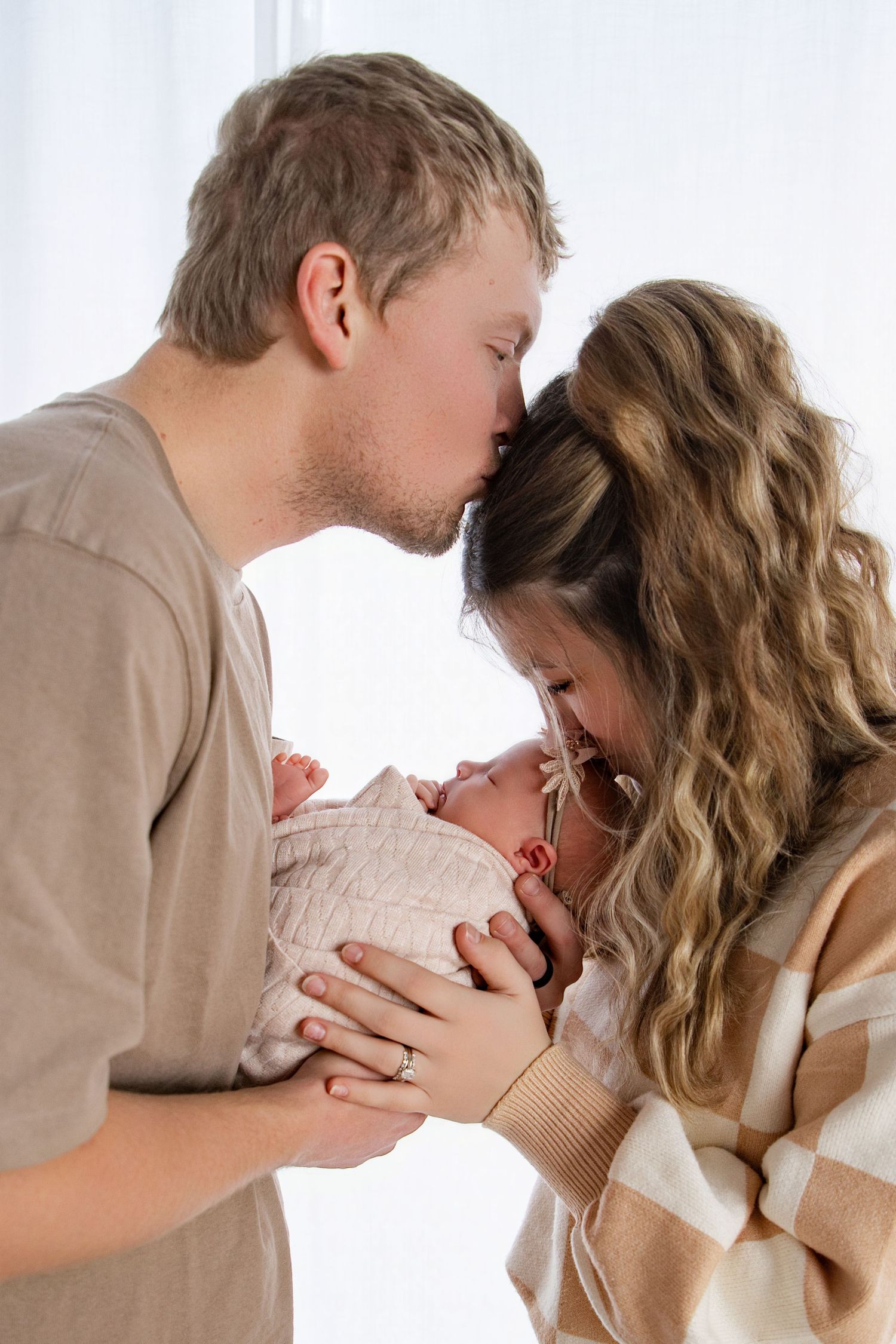 A couple shares a tender kiss over their newborn baby, wrapped in a white swaddle, in a bright softly lit room.