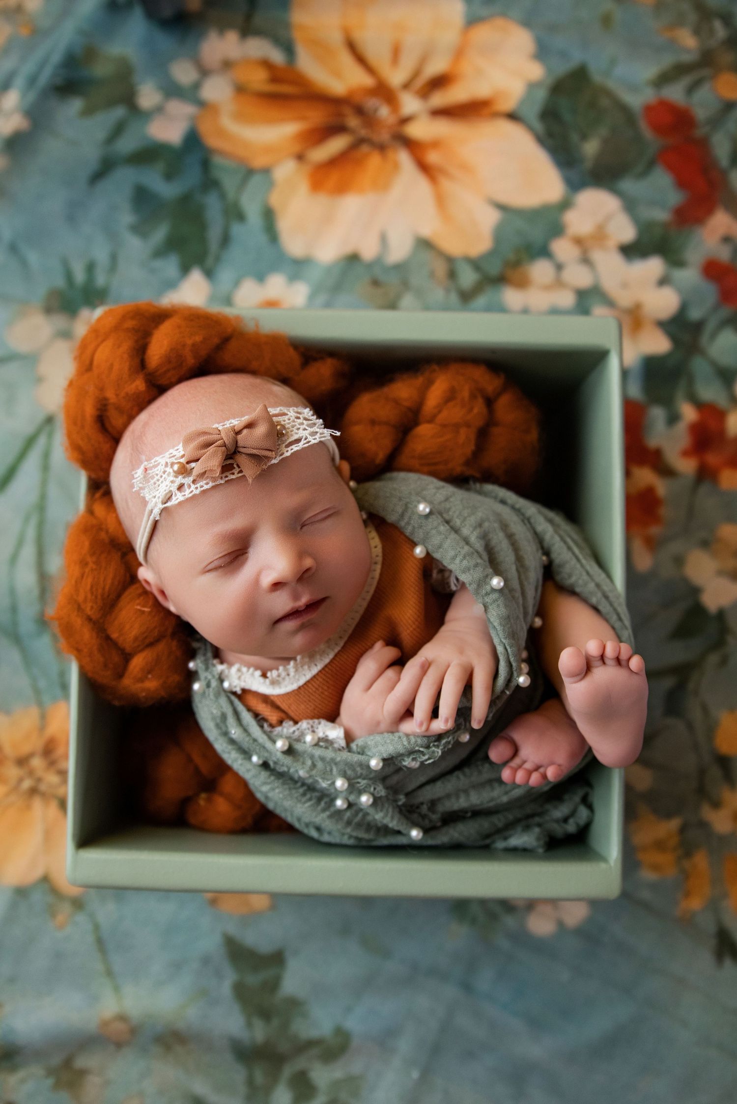 Sleeping newborn baby wrapped in sage green cloth with pearl details, wearing a lace headband, nestled in a mint green box.