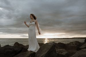 A figure in a white wedding dress stands gracefully on coastal rocks at sunset under dramatic stormy clouds.