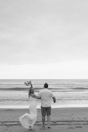 A couple stands on a beach in black and white, with the bride's dress flowing in the wind as they face the ocean.
