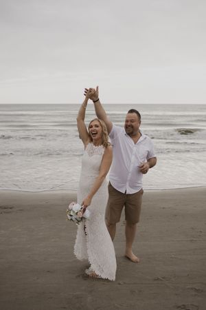 Couple dancing joyfully on a misty beach with bride in white lace dress and groom in casual attire.