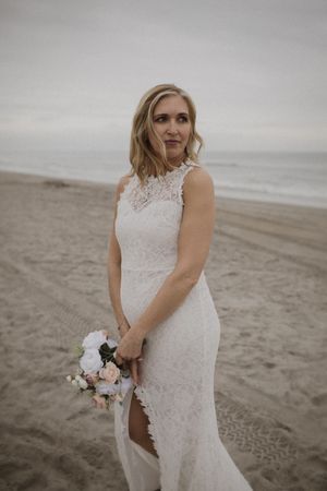 A bride in a lace wedding dress poses on a cloudy beach holding a small bouquet of pink and white flowers.