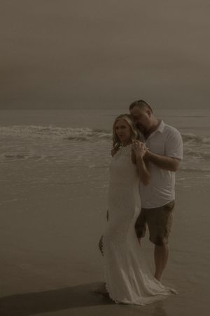 A couple in wedding attire shares an intimate moment together on a misty beach at dusk.