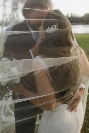 A romantic wedding moment captured through a flowing veil as a couple embraces outdoors near water.