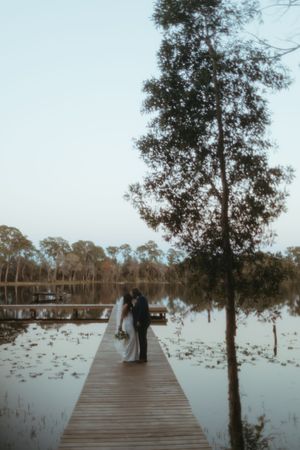 A couple stands on a wooden boardwalk over a lily-covered lake surrounded by eucalyptus trees at dusk.