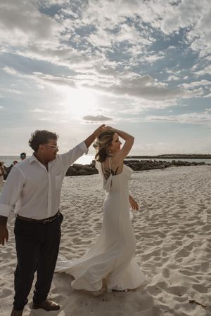 A couple dances playfully on a sandy beach at sunset with dramatic cloudy skies overhead.