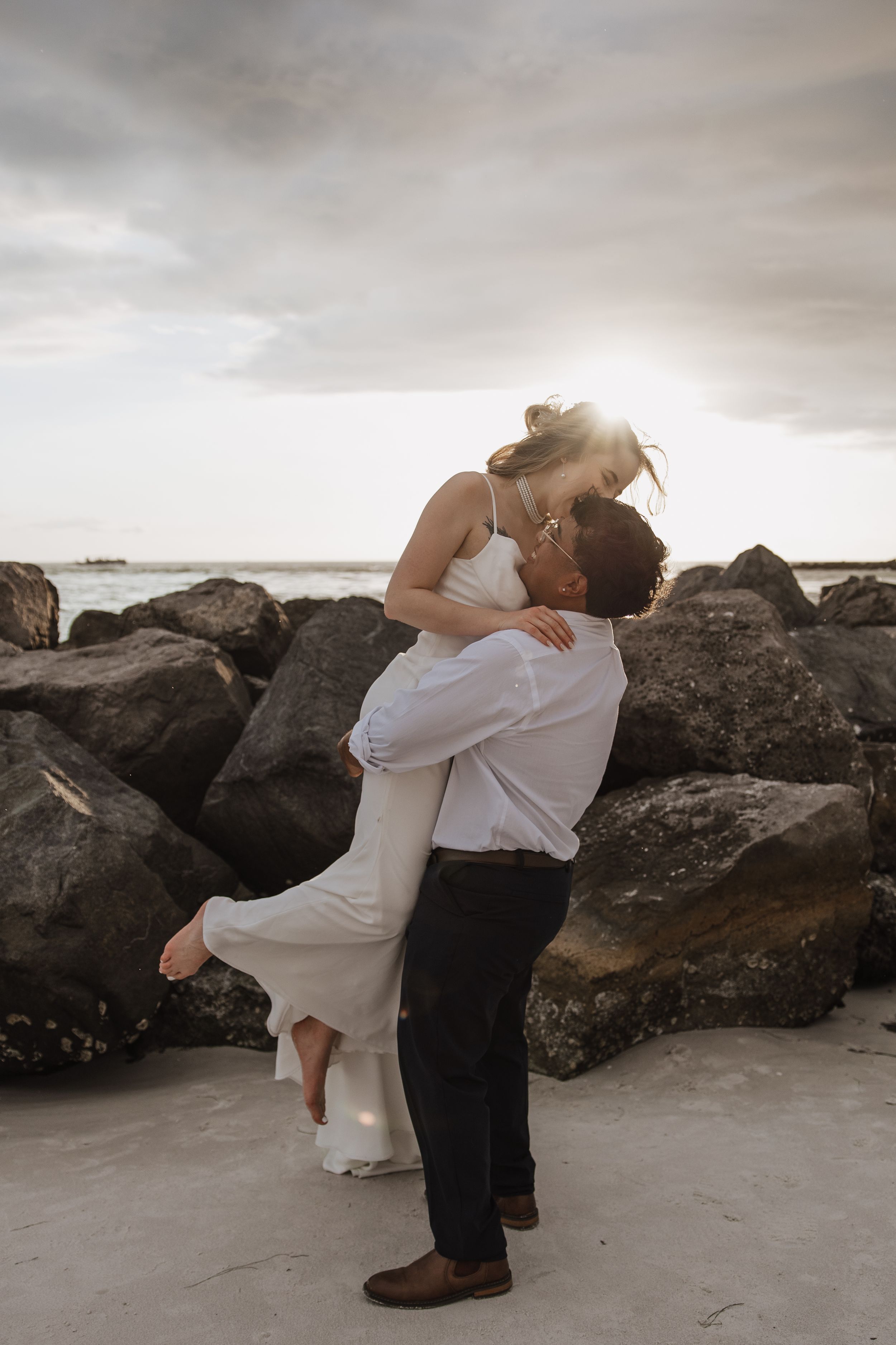 A romantic couple embraces on a rocky beach under moody cloudy skies during a wedding photoshoot.