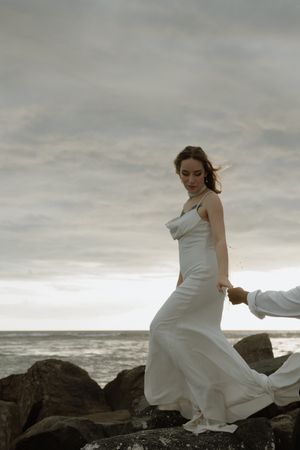 Dramatic seaside wedding photos with bride in flowing white dress against rocky coastal backdrop and moody sky.