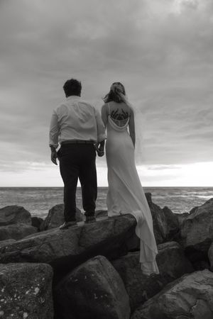 A couple stands together on rocky coastal outcropping overlooking stormy ocean in black and white photograph.