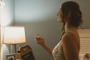 A person in a white dress holds a glass while standing in a softly lit room with a table lamp visible.