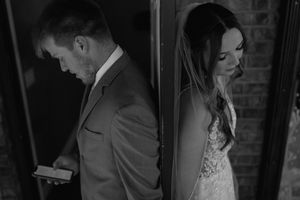 A bride and groom stand back to back against a wall in a dramatic black and white wedding photo.