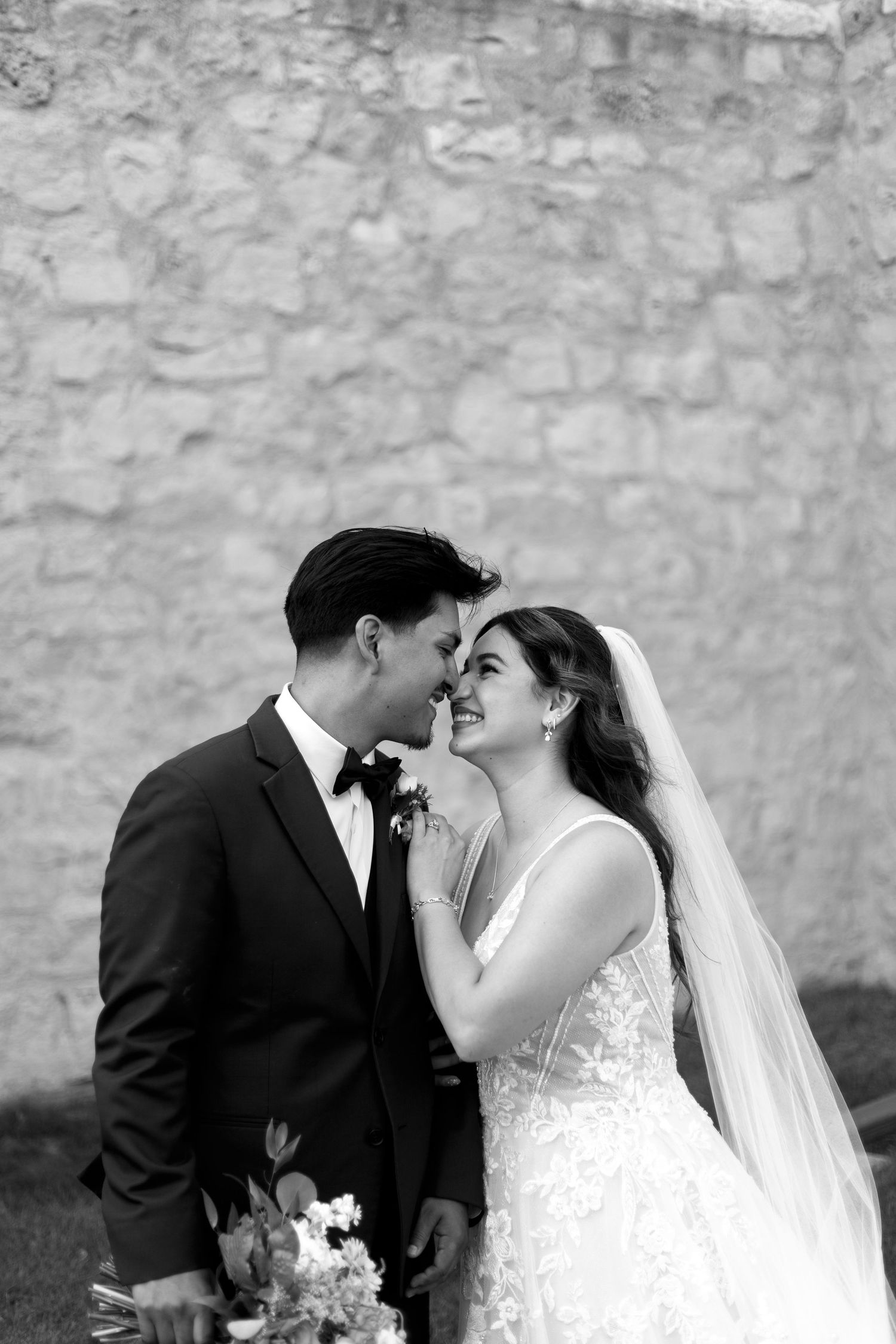 A romantic black and white wedding portrait of a couple sharing an intimate moment against a stone wall.