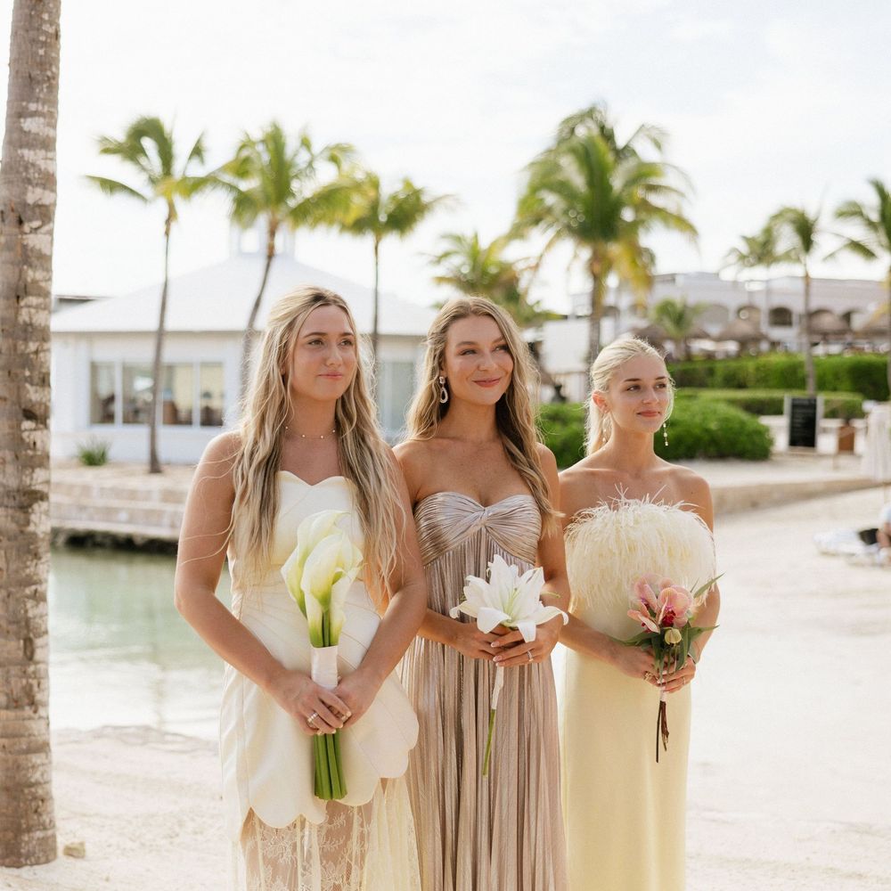 Three bridesmaids in light-colored dresses hold floral bouquets while standing on a tropical beach with palm trees.