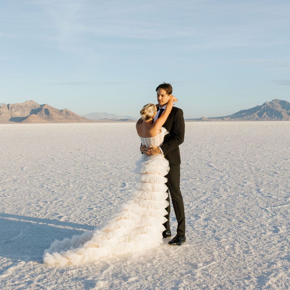 A couple embraces on the salt flats during their wedding photoshoot with mountains in the background at sunset.