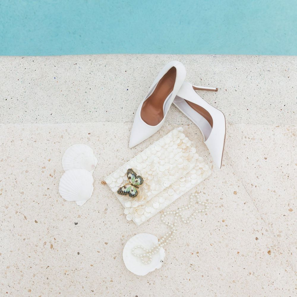 White stiletto heels with wedding accessories including pearl jewelry and delicate lace details on light beige surface.