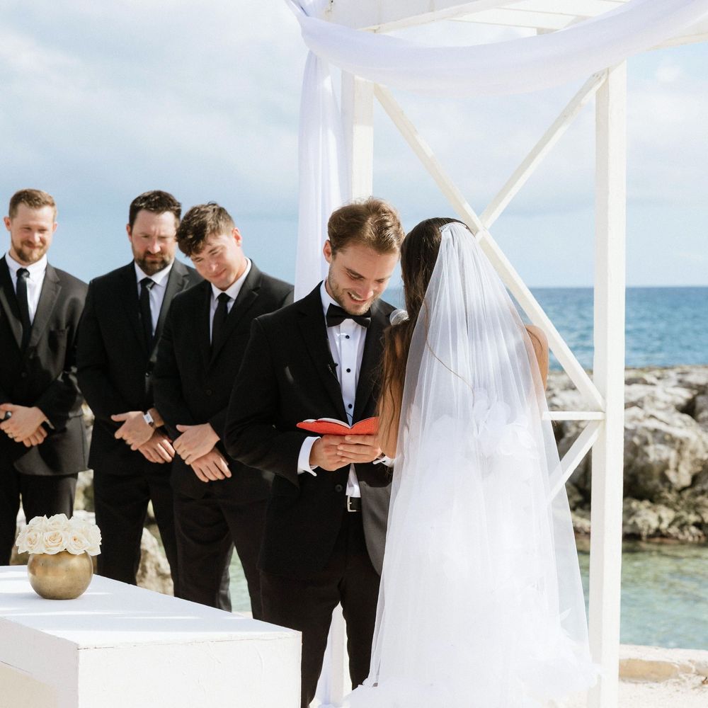 A wedding party in black tuxedos and a bride in white share a joyful moment together by the ocean.